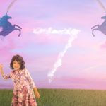 Girl in unicorn dress holding a pink water balloon with a pink sky behind her. In the pink sky are clouds forming the number seven and two unicorns with rainbows coming out of their horns. Orlando children's portraits.