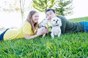 Husband and wife laying on the ground with their dogs as one dog kisses husband while husband makes a silly kiss face.